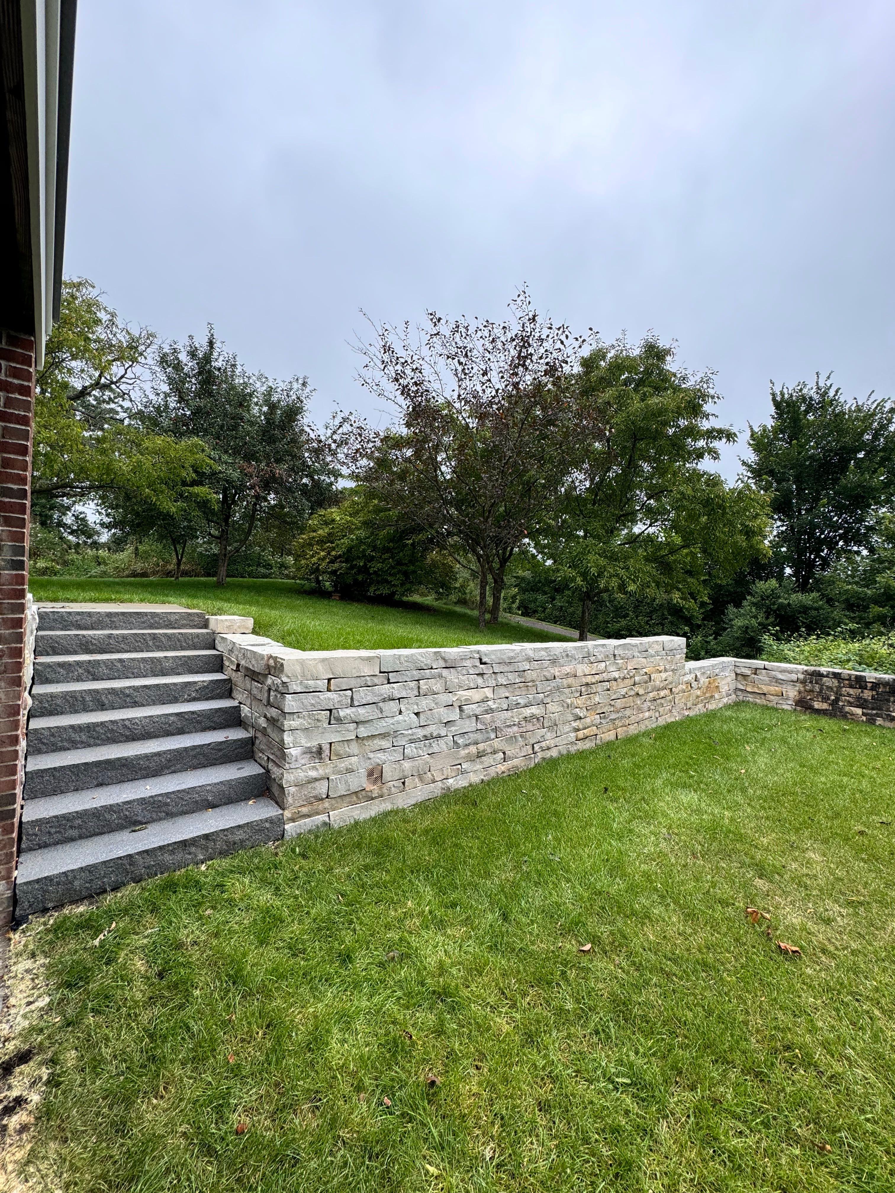 Stone steps leading to a landscaped yard with trees and a stone wall under a cloudy sky.