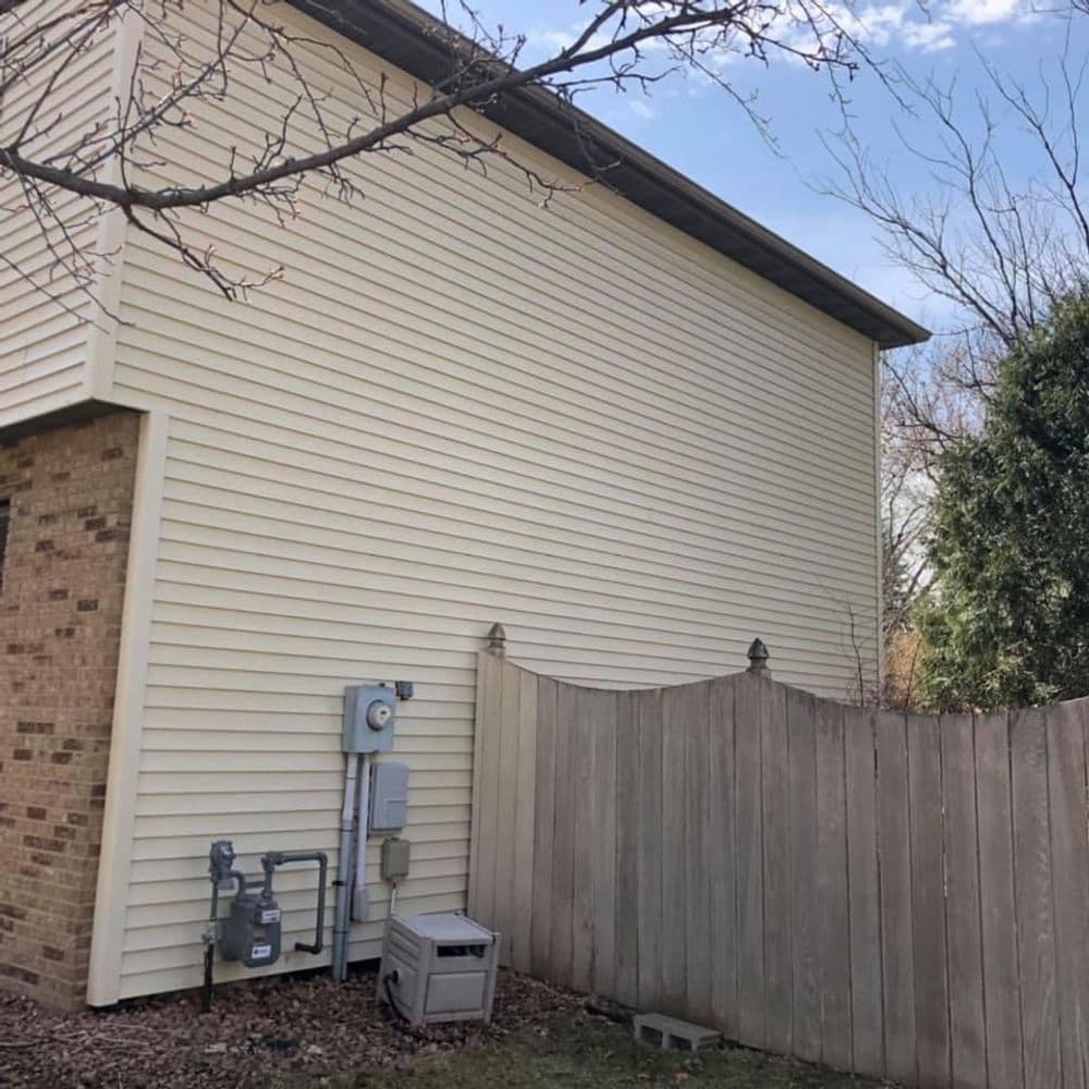 Side view of a residential house with beige siding and a wooden fence. Gas meter visible.